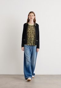 Zwarte blazer met voering in luipaardprint, groene gebloemde top, en wijde blauwe jeans. Model staat op een effen achtergrond.