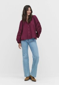 Frau mit langen dunklen Haaren, die eine lockere lila Bluse mit Spitzenärmeln, hellblaue Jeans und braune Absatzstiefel trägt, vor einem weißen Hintergrund steht.