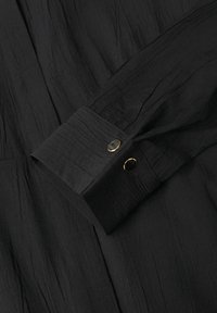 Dettaglio di una camicia nera con un tessuto testurizzato, un polsino piegato con bottoni dorati e neri e linee di cucitura visibili.