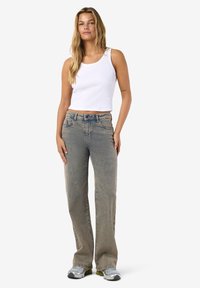 Jauna sieviete stāv, tērpusies baltā bezroku crop topā, izbalējušās taisnajās džinsu biksēs un sudraba krāsas sporta apavos, uz vienkārša fona.