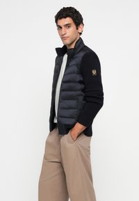 Homme portant une veste matelassée et tricotée noire, une chemise blanche et un pantalon beige, les mains dans les poches, se tenant devant un fond blanc uni.
