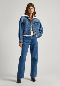 Veste en denim bleu avec col blanc, portée avec un haut en tricot blanc et un jean en denim. Les bottes sont noires. Coupe droite, design décontracté.