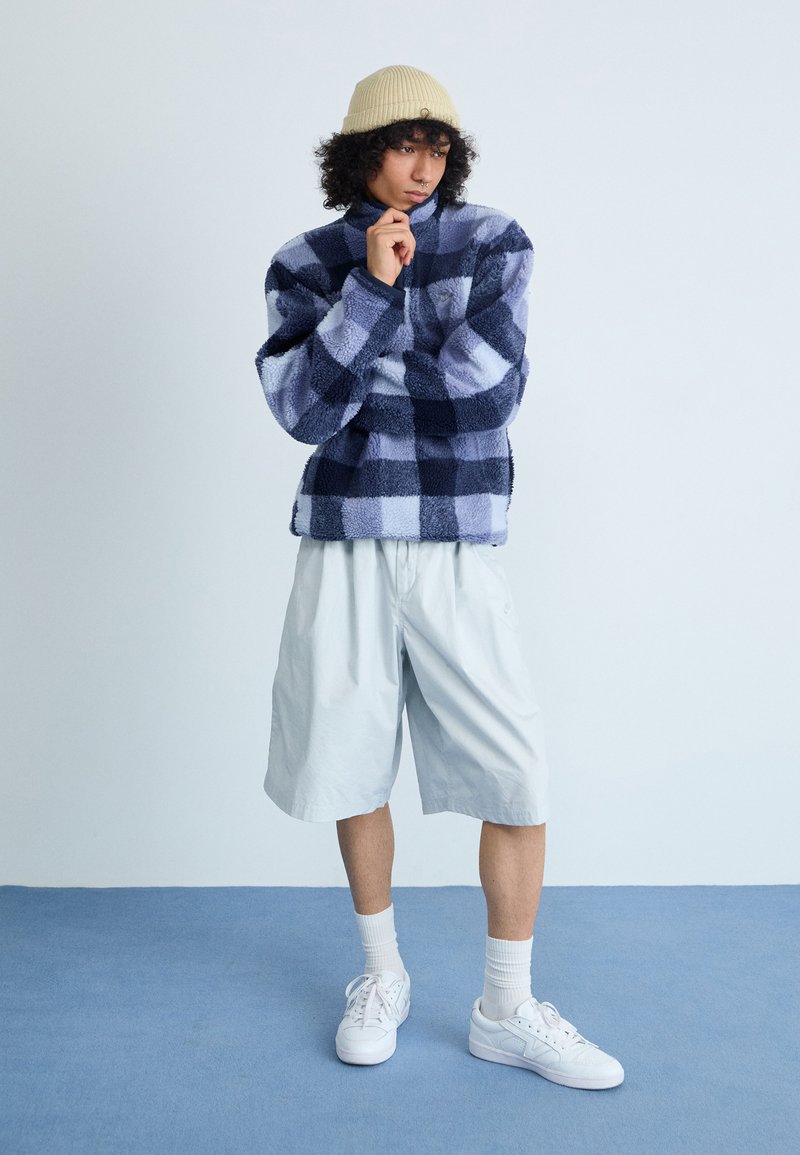 Pullover in pile con motivo a quadri navy e azzurro, pantaloni corti chiari, sneakers bianche e berretto di maglia beige, in piedi su uno sfondo blu.