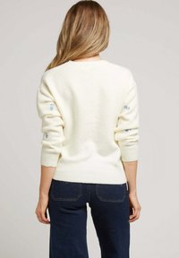 Un pull de couleur crème en matériau doux avec des poignets roulés et des broderies florales bleues sur les manches. Porté avec un jean bleu foncé.