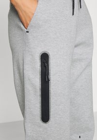 Grå sweatpants gjorda av ett mjukt material har en svart dragkedjeficka på låret med en texturerad detalj. Dragsko-stängning vid midjan.