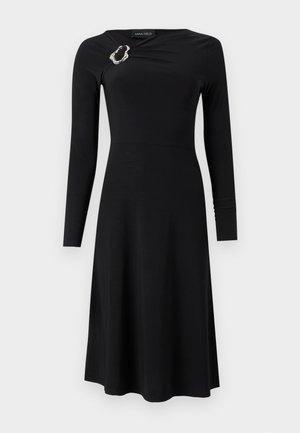 Robe midi noire à manches longues avec encolure ronde et détail asymétrique d'une broche argentée sur l'épaule gauche.