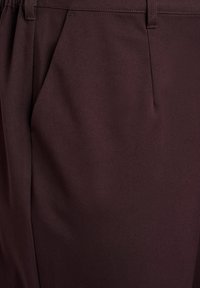 Bordeauxrode broek van gladde stof, met een geplooid voorpand, zijzakken en een klassieke tailleband met riemlussen.