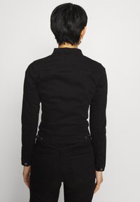 Chaqueta de mezclilla negra con una forma ajustada, mangas largas y puños con botones. Presenta detalles en la parte posterior y una textura suave.