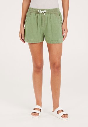 Shorts de lino verdes con cintura elástica, detalle de cordón, bolsillos laterales y un corte relajado. Combinados con sandalias blancas.