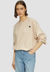 Camicia beige a righe oversize con maniche arrotolate e un piccolo emblema a forma di cuore nero sul lato sinistro del petto, abbinata a jeans di denim blu chiaro.
