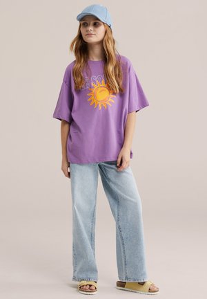 Giovane che indossa una t-shirt viola oversize con una grafica del sole, jeans a gamba larga azzurri chiaro, sandali beige e un berretto da baseball azzurro chiaro.