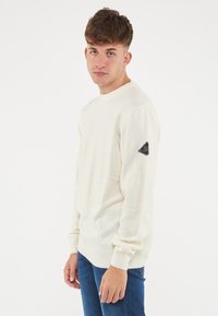 Maglione color crema con collo rotondo, polsini a coste e patch con logo sulla spalla sinistra. Realizzato in un materiale morbido e leggero.