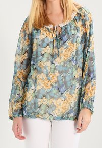 Halvgennemsigtig bluse med blomstertryk, der har blå, grønne og gule blomster. Snoretræk i halsen og elastiske manchetter. Blødt, let materiale.