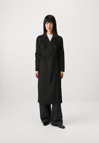 Cappotto lungo nero con cintura, dotato di colletto a revers e maniche lunghe. Abbinato a jeans di denim scuri a gamba larga e scarpe nere.