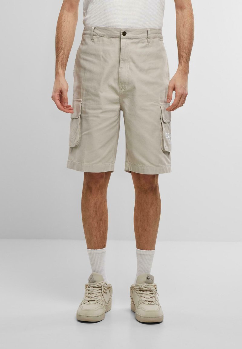 Pantaloni cargo beige con tasche laterali, realizzati in cotone. Presentano una chiusura con bottone e una vestibilità dritta, abbinati a scarpe da ginnastica chiare e calze bianche.