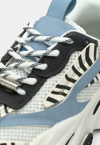 Baskets avec une tige en mesh blanche, des accents rayés noirs, des superpositions bleu clair et des lacets texturés avec un œillet noir et une étiquette.