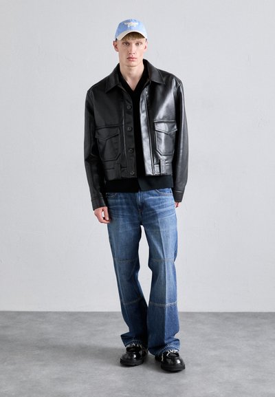 Czarna skórzana krótka kurtka z dwiema przednimi kieszeniami, zestawiona z luźnymi niebieskimi dżinsami. Model nosi jasnoniebieską czapkę i czarne buty.