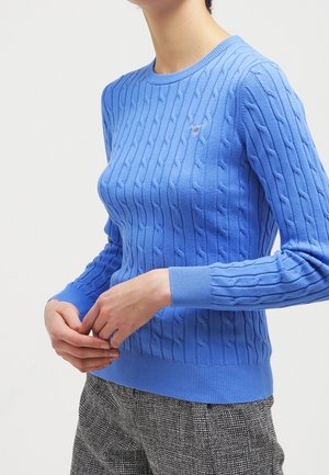 Personne portant un pull en maille torsadée bleu ajusté avec poignets et ourlet côtelés, associé à un pantalon gris à carreaux, mains légèrement jointes.