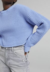 Personne portant un pull en tricot bleu clair à manches longues et un jean gris d�élavé, la main glissée dans la poche avant, sur fond uni.
