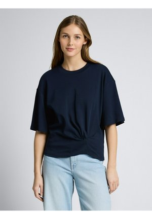 Młoda kobieta o długich, jasnobrązowych włosach, ubrana w luźną, ciemnoniebieską bluzkę z krótkim rękawem i jasnoniebieskie dżinsy, stojąca na jednolitym tle.