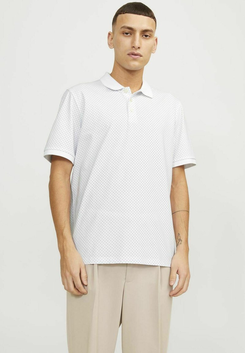 Polo bianco con un motivo a puntini discreto, maniche corte, colletto e patta con due bottoni. Abbinato a pantaloni beige.