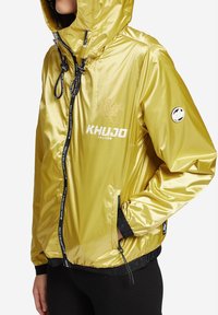 Žlutá, lesklá bunda na zip s kapucí, šňůrkami, předními kapsami a logem. Vyrobena z hladkého, lehkého materiálu.