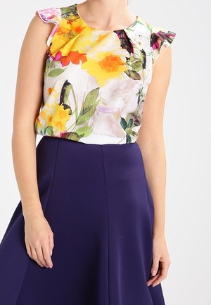 Bezrekawnik z żółto-różowym kwiatowym wzorem w połączeniu z wysokim stanem i ciemnofioletową spódnicą w kształcie litery A na stojącej osobie.