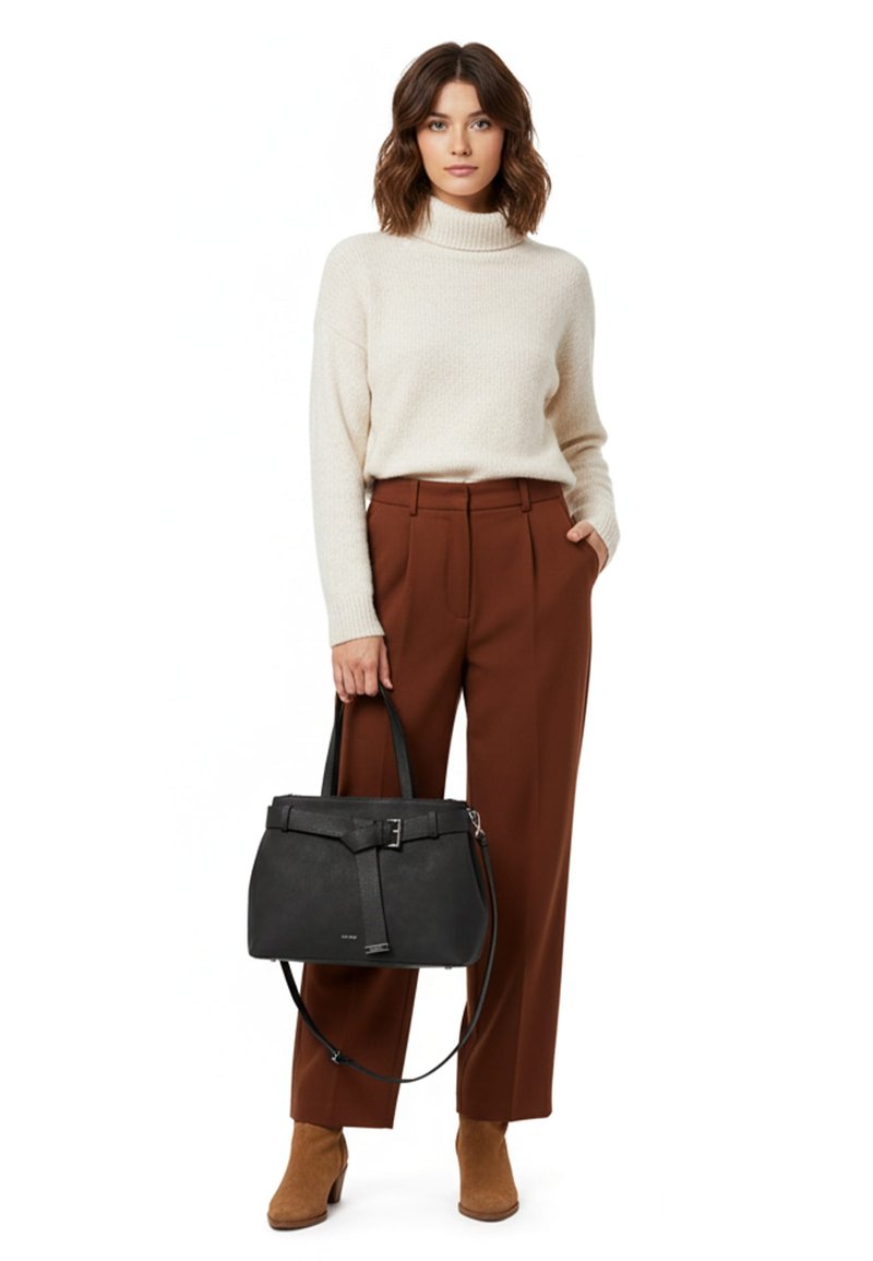 Naine, kes kannab kreemikat kõrge kaelusega kampsunit ja roostepunaseid pükse, hoides käes musta käekotti, seljas pruunid pahkluu saapad valgel taustal.