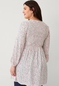 Bloes met bloemenmotief en een gesmokte rug, versierd met roze bloesems en groene bladeren op een witte achtergrond. Lange, opgezette mouwen maken het ontwerp compleet.