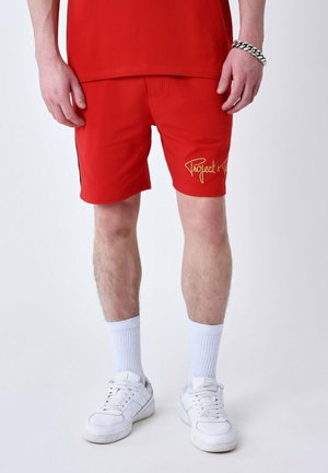 Pantalones cortos rojos con una franja lateral negra y "Projecto" en letras doradas en la pierna izquierda, combinados con calcetines blancos y zapatillas.