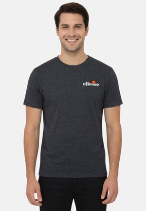 Homme souriant portant un t-shirt ellesse gris foncé et un pantalon noir, debout devant un fond clair uni.