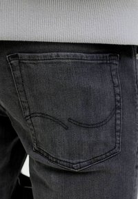 Black denim jeans with a back pocket featuring a subtle curved seam design. The fabric has a smooth, textured appearance.