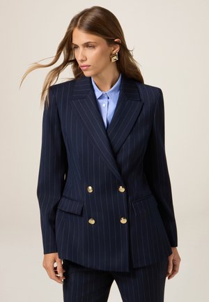 Blazer azul marino de doble botonadura con botones dorados y rayas finas. Corte ajustado, solapas con muesca y dos bolsillos delanteros. Camisa azul clara debajo.