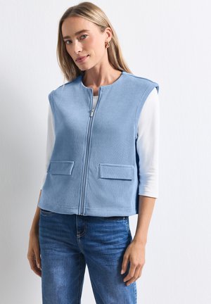 Blonde vrouw die een lichtblauw mouwloos vest met rits draagt over een wit shirt met lange mouwen en een blauwe spijkerbroek, staand tegen een effen witte achtergrond.