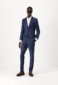 Navy blue suit jacket and trousers with a subtle check pattern, paired with a white shirt. Brown leather shoes and dark sunglasses.