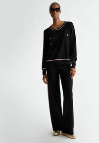 Maglione nero a maglia con rifiniture dorate e due taschini sul petto, abbinato a pantaloni neri a gamba larga con una striscia dorata sui lati. Scarpe dorate.