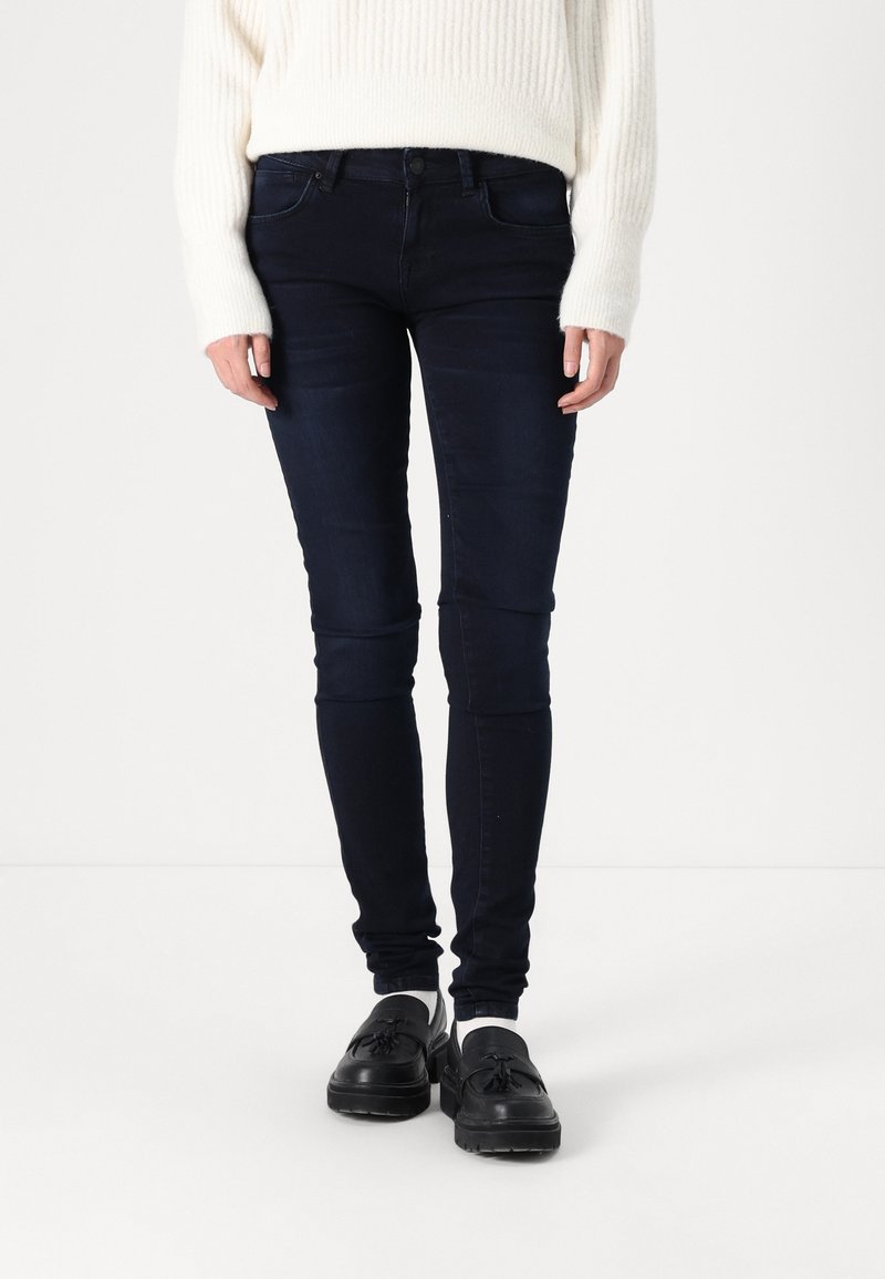 Personne portant un jean skinny foncé, un pull blanc, des chaussettes blanches et des mocassins noirs à pampilles, debout sur un sol et un fond blancs.