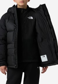Geacă matlasată neagră cu fermoar și căptușeală interioară din fleece, având logo-ul The North Face pe piept și o etichetă pe tiv.