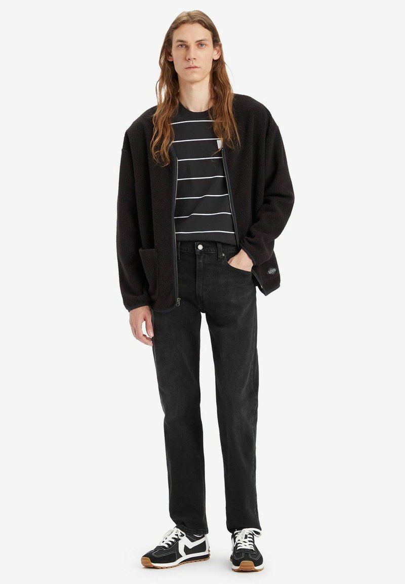 Schwarze Fleece-Jacke mit Reißverschluss und Seitentaschen über einem schwarz-weiß gestreiften T-Shirt, kombiniert mit schwarzen schmal geschnittenen Jeans und schwarzen Sneakers.