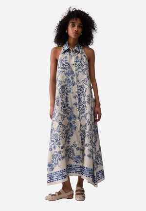 Mujer con cabello rizado, vestida con un vestido sin mangas color beige con estampado floral azul y sandalias beige con tachuelas, de pie sobre un fondo blanco.