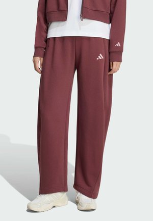 Persona con pantalones anchos de chándal Adidas granate y chaqueta a juego con zapatillas blancas, de pie frente a un fondo liso.