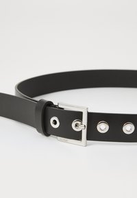 Černý kožený opasek s elegantním designem, který je vybaven stříbrnou obdélníkovou sponou a kovovými očky podél pásu. Logo je ražené na sponě.