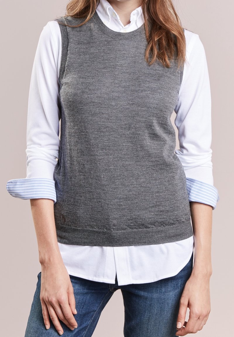 Frau, die ein weißes Hemd mit aufgerollten Ärmeln unter einem ärmellosen grauen Pullover und blauen Jeans trägt, vor einem einfarbigen Hintergrund steht.