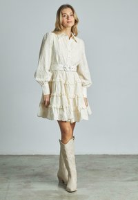 Cremiges Spitzenkleid mit Rüschenschichten, langen Ärmeln und tailliertem Bund, kombiniert mit knielangen beigefarbenen Cowboy-Stiefeln auf einem Betonboden.