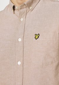Lichtbruine button-up shirt met een gestructureerde stof, voorzien van een enkele borstzak en een klein zwart en geel adelaarslogo.