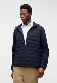 Νεαρός άντρας που φοράει ναυτικό μπουφάν με φερμουάρ και καπιτονέ επένδυση πάνω από ανοιχτό μπλε πουκάμισο και μπεζ παντελόνι, στέκεται μπροστά σε απλό φόντο.