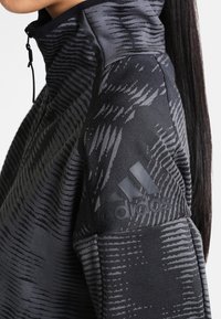 Veste de sport noire avec un col haut, motif texturé et fermeture éclair au cou. Présente un logo noir sur l'épaule. Tissu doux.