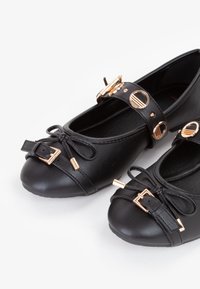 Ballerine sintetiche nere con punta tondeggiante, dettagli in metallo dorato, fiocco decorativo e una fascia con ritagli circolari.