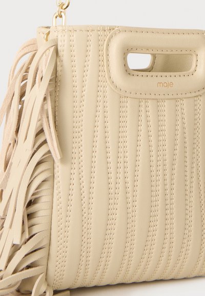 Sac à main en cuir beige avec coutures verticales, anse découpée, métal doré, et longs pompons à franges sur le côté.