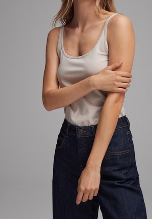 Kobieta ubrana w białą bluzkę na ramiączkach i ciemnoniebieskie dżinsy, z jedną ręką skrzyżowaną i trzymająca przeciwległe ramię na jednolitym szarym tle.